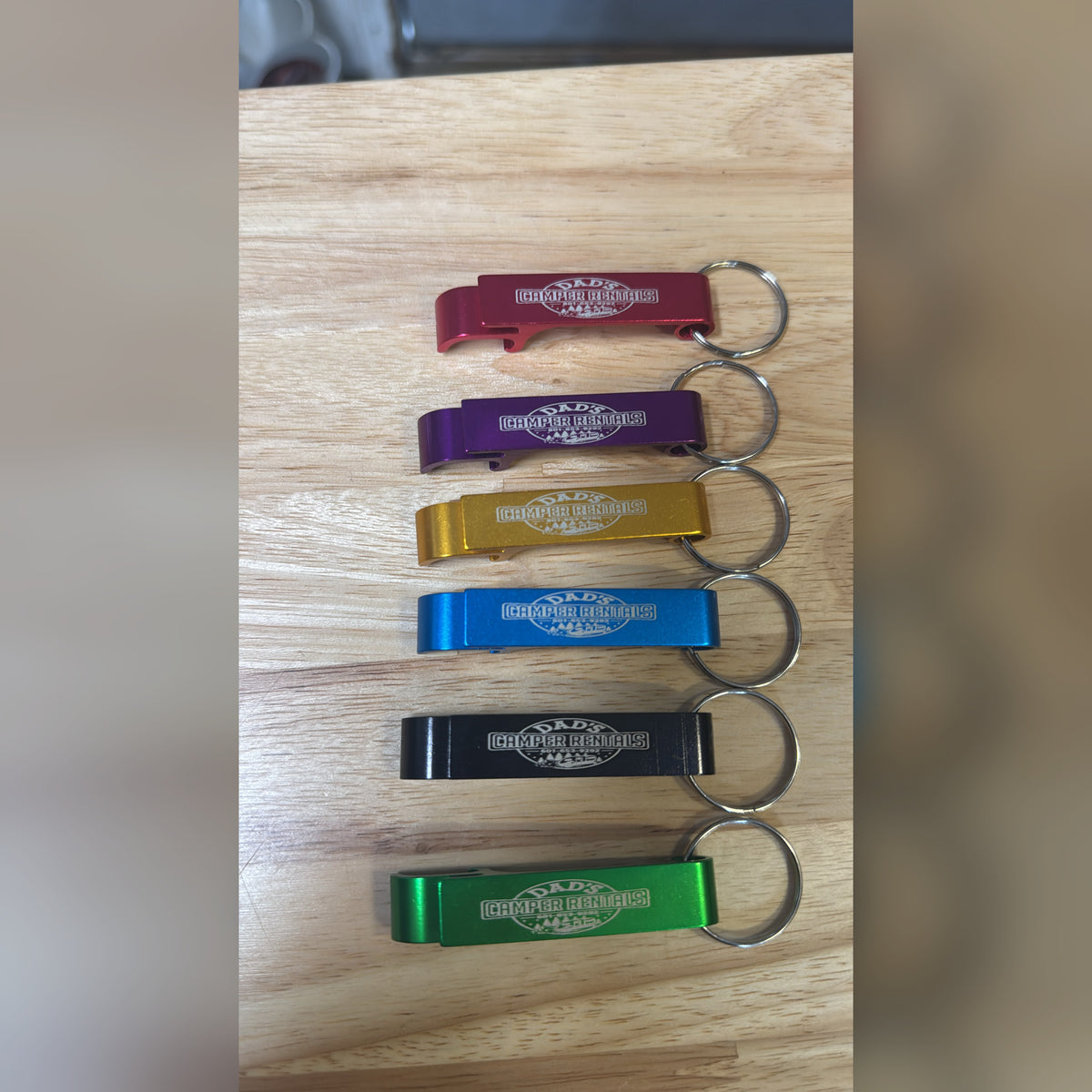 Six custom laser-engraved metal bottle openers in red, purple, gold, blue, black and green lined up on a wood table